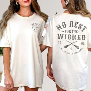 May include: Cream-colored t-shirt with the text "NO REST FOR THE WICKED" in dark gray. The shirt features a small graphic on the front and a larger design on the back, including crossed brooms and the words "DAUGHTERS OF MIDNIGHT."