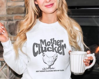 Może przedstawiać: Biała bluza z czarnym nadrukiem kurczaka i tekstem "Mother Cluckin' Ain't Nobody Cluckin' With My Flock"