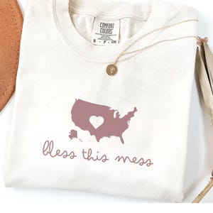 May include: A white t-shirt with a pink outline of the United States with a heart in the middle. The text "bless this mess" is printed below the map.