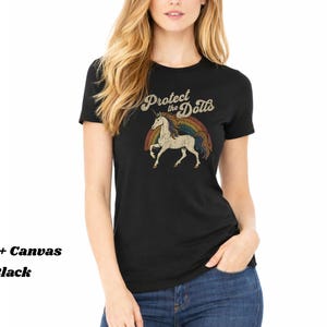 May include: Black t-shirt with a vintage-style graphic featuring a unicorn, rainbow, and the text "Protect the Dolls." The shirt is made of Bella + Canvas material.