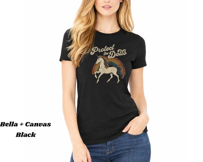 May include: Black t-shirt with a vintage-style graphic featuring a unicorn, rainbow, and the text "Protect the Dolls." The shirt is made of Bella + Canvas material.