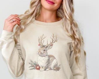 Puede incluir: Sudadera beige con una ilustración en acuarela de un conejo con cuernos de ciervo, adornada con adornos navideños. El diseño incluye agujas de pino y bayas rojas, creando un tema festivo navideño.