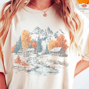 Può includere: T-shirt color crema con un paesaggio montano panoramico con baite, alberi e un ruscello. La grafica include colori autunnali come arancione e giallo, con un tema rustico e all'aperto. La maglietta ha una vestibilità ampia.