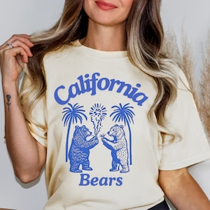 May include: Cream-colored t-shirt with a blue graphic featuring the words "California Bears." The design includes two bears, palm trees, and fireworks. The shirt is a relaxed fit.