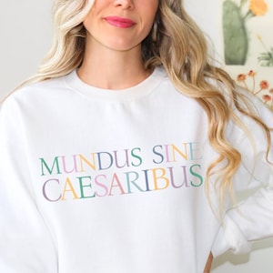 Könnte beinhalten: Weißer Pullover mit dem Text "MUNDUS SINE CAESARIBUS" in bunten Buchstaben.