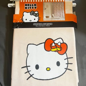May include: A Hello Kitty shower curtain, 72 x 72 inches (183cm x 183cm), featuring a large graphic of Hello Kitty with a candy corn bow. The packaging includes the Hello Kitty logo and a picture of the curtain in use.