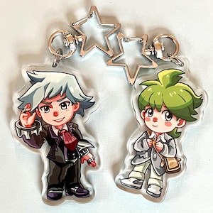 May include: Two clear acrylic keychains featuring anime-style illustrations of male characters. One character has white hair and wears a black suit with a red tie. The other has green hair and a gray jacket. Both have silver star-shaped clasps.