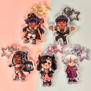 May include: Five acrylic keychains featuring cartoon characters with star-shaped charms. The characters are depicted in various outfits and poses, with a pink and blue background. Each keychain has a metal clasp.