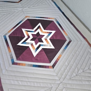 May include: Close-up of a handmade quilt with a geometric star design. The quilt features a central star in shades of burgundy, white, and orange, set within a hexagonal shape. The background is a light gray with stitched lines.