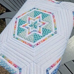 May include: A white quilted blanket with a central star design. The star is composed of turquoise and rainbow-colored polka dot fabric, surrounded by a white hexagon. The blanket features a geometric pattern with white and rainbow accents.