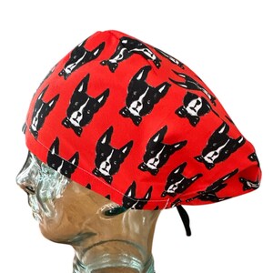 May include: A red surgical scrub cap featuring a repeating pattern of black and white Boston Terrier dog faces. The cap has a tie in the back for an adjustable fit. The cap is made of a soft, lightweight fabric.