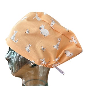 May include: Peach-colored surgical scrub cap with a repeating pattern of white cartoon animals, including dogs, pigs, and birds. The cap has a gathered back and a white drawstring for adjustment. The fabric is a soft cotton.