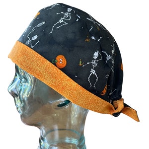 Könnte beinhalten: Eine schwarze OP-Mütze mit orangefarbenem Besatz. Die Mütze hat ein Muster aus Skeletten und Kürbissen.