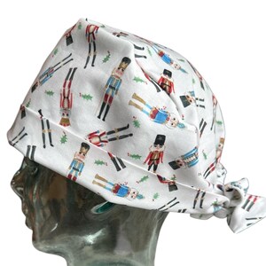 Op de afbeelding: Witte stoffen muts met een herhalend patroon van kleurrijke notenkrakerfiguren en groene hulstbladeren. De muts is aan de achterkant vastgebonden en wordt getoond op een heldere glazen hoofdvorm.