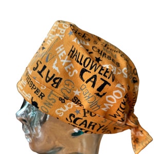 Op de afbeelding: Oranje scrub-hoed met een Halloween-thema print met woorden als "Halloween", "Kat", "Vleermuizen", "Wicked", "Eng", "Vloeken", "Hexen", "Fluisteren", "Spookachtig", "Leuk", "Maan" en "Trick".
