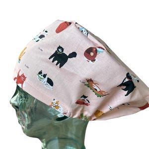 Pode incluir: Uma touca cirúrgica rosa claro com um padrão repetido de gatos caprichosos em várias poses e cores. Os gatos são retratados em preto, branco, laranja e tartaruga, alguns com acessórios. A touca é projetada para profissionais médicos.