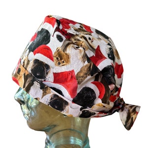 Könnte beinhalten: Eine OP-Haube mit einem Muster aus Hunden mit Weihnachtsmützen. Die Haube ist weiß und rot und wird am Rücken gebunden. Sie wird auf einem Glaskopf präsentiert.