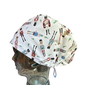 May include: White surgical scrub cap with a repeating pattern of toy nutcracker soldiers in red, blue, and gold. The cap has a drawstring and is designed to cover the hair during medical procedures.