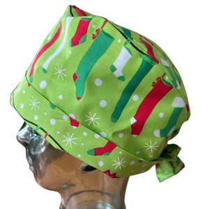 以下が含まれることがあります： 緑色のクリスマスストッキング柄のスクラブキャップ。赤、緑、白のストッキングと雪の結晶が薄緑色の背景に描かれています。キャップは後ろで結ばれています。