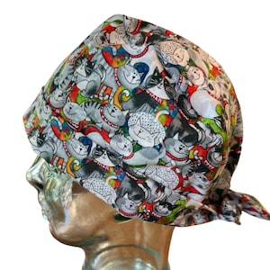 May include: A white surgical scrub hat with a colourful print of cats. The cats are in various poses and have different expressions. The hat has a tie at the back.