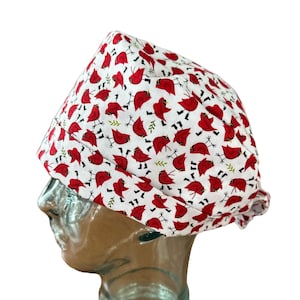 May include: A white cap featuring a repeating pattern of red cardinal birds and green sprigs. The cap is made of fabric and is designed to be worn on the head. The cap is shown on a mannequin head.