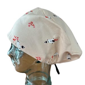 May include: A light beige surgical scrub cap featuring a pattern of raccoons and red floral designs. The cap has a black drawstring. The fabric appears to be cotton. The cap is designed for comfort.