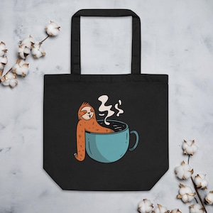 May include: Black tote bag featuring a cartoon sloth relaxing in a blue coffee cup. The sloth is orange with a smiling face, and steam rises from the coffee. The bag has two black handles and is made of a sturdy material.