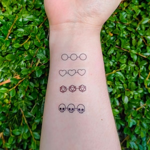 Op de afbeelding: Een pols met vier tijdelijke tattoo-ontwerpen: brillen, harten, dobbelstenen en schedels. De tatoeages zijn zwart-wit, met een minimalistische stijl. De achtergrond is een groene, bladerrijke plant.