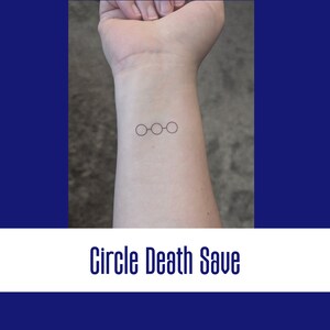 Op de afbeelding: Een close-up van iemands pols met een kleine tatoeage van drie verbonden cirkels. De tatoeage is een eenvoudige lijntekening en bevindt zich aan de binnenkant van de pols. De tatoeage wordt "Circle Death Save" genoemd.