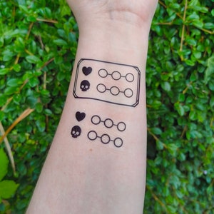 May include: Black temporary tattoos applied to the skin. The designs include a heart, a skull, and a series of connected circles. The tattoos are displayed on a person's arm against a background of green foliage.