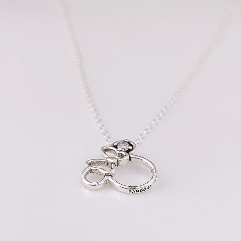 K&ouml;nnte beinhalten: Eine silberfarbene Halskette mit einem Anh&auml;nger in Form einer stilisierten Schleife und eines Rings. Der Anh&auml;nger hat einen kleinen, klaren Edelstein und das Wort "Pandora" ist auf dem Ring eingraviert. Die Kette ist ebenfalls silberfarben.