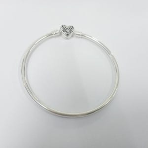 Puede incluir: Una pulsera de brazalete plateada con un dije en forma de corazón. El dije tiene un diseño calado detallado. La pulsera es circular y parece estar hecha de un metal brillante, posiblemente plata de ley.