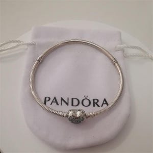 May include: A silver bracelet with a heart-shaped charm, resting on a white pouch. The bracelet has a snake chain design and a heart charm with small, sparkling accents. The pouch has the brand name "Pandora" printed on it.