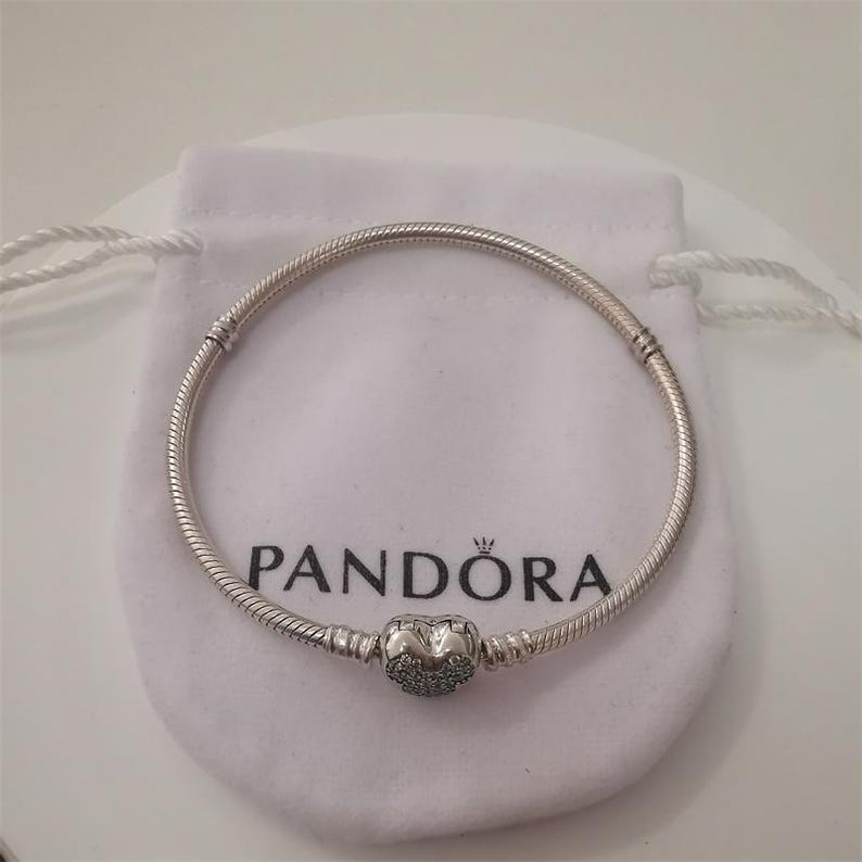 May include: A silver bracelet with a heart-shaped charm, resting on a white pouch. The bracelet has a snake chain design and a heart charm with small, sparkling accents. The pouch has the brand name "Pandora" printed on it.