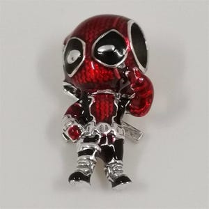 May include: A red and black Deadpool charm with silver accents. The charm features a detailed design of the character in a standing pose, with a textured finish on the red areas. The charm is likely made of metal and is designed to be attached to a bracelet or necklace.