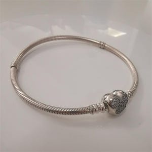May include: A silver-colored bracelet with a snake chain design. The bracelet features a heart-shaped clasp adorned with small, sparkling stones. The bracelet is a piece of jewelry.