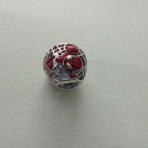 May include: A round, silver charm with a red and black Spider-Man design. The charm features a webbed sphere with a red Spider-Man figure inside. The design is detailed and eye-catching, suitable for a bracelet or necklace.