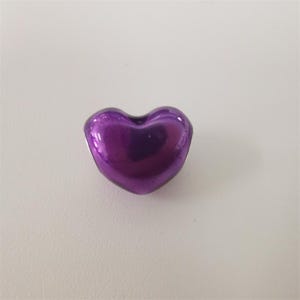 May include: A small, metallic purple heart-shaped object. The heart has a smooth, reflective surface, and is set against a white background. Likely a decorative item or charm.
