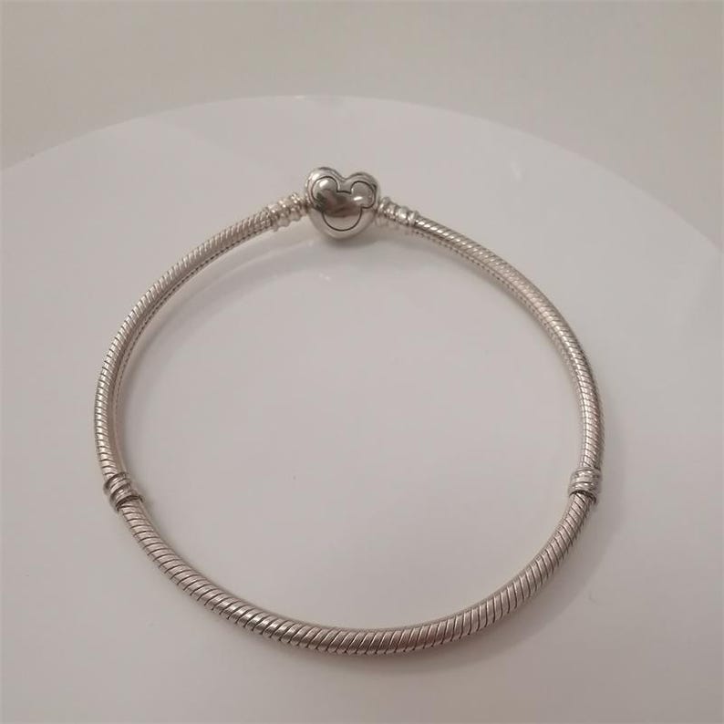 May include: A silver bracelet with a heart-shaped clasp featuring a Mickey Mouse design. The bracelet has a snake chain design and is approximately 7 inches in length. The clasp is detailed with a small Mickey Mouse face.