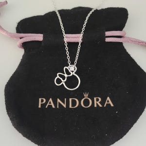 K&ouml;nnte beinhalten: Eine silberne Halskette mit einem anh&auml;nger in Schleifenform mit einem kleinen, funkelnden Edelstein. Die Halskette liegt auf einem schwarzen Samtbeutel mit dem goldfarbenen Aufdruck "PANDORA". Der Beutel hat einen rosa Kordelzug.