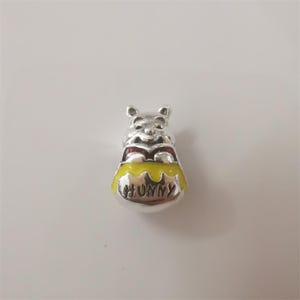 May include: A silver charm depicting Winnie the Pooh, holding a yellow honey pot. The word "HUNNY" is written on the pot. The charm has red and yellow enamel detailing.