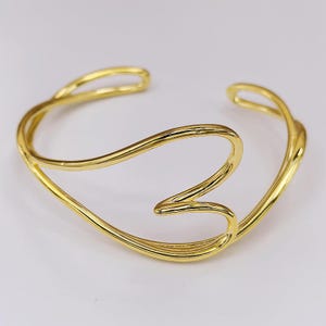 May include: A gold-colored cuff bracelet with a flowing, open design. The bracelet features a continuous, curved line that forms an open shape. The metal has a polished, reflective surface, and the overall style is modern and elegant.