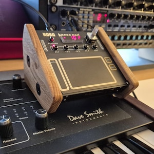 以下が含まれることがあります： 木製のフレームが付いたKorg Kaoss Pad 4+電子楽器。Dave Smith Instrumentsシンセサイザーの上に置かれています。 Kaoss Padには黒いタッチパッド、ディスプレイ、さまざまなコントロールがあります。シンセサイザーにはノブとキーがあります。