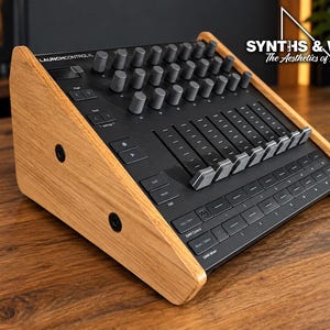 May include: A black Launch Control XL MIDI controller housed in a light-coloured wooden frame with side panels. The device features multiple grey rotary knobs, eight vertical faders, and various buttons. The text SYNTHS & WOOD The Aesthetics of Sound is visible in the upper right corner.