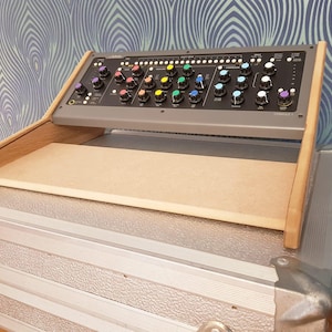 Peut inclure: Un support en bois avec une surface supérieure beige, conçu pour accueillir une console de mixage audio. La console a une face noire avec des rangées de boutons et de molettes, certains avec des accents colorés.