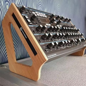 Puede incluir: Un soporte de madera para un sintetizador Novation Summit. El soporte está hecho de madera marrón claro y tiene una forma triangular. El sintetizador es negro con detalles azules y tiene el texto "Summit Analog Synthesizer" impreso en la parte delantera.