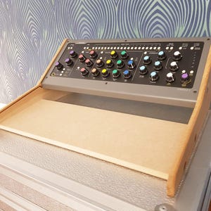 May include: A wooden stand with a beige top surface, designed to hold a mixing console. The console has a gray metal casing with a black control panel featuring colorful knobs and buttons. The stand is angled to provide a comfortable viewing angle for the console.