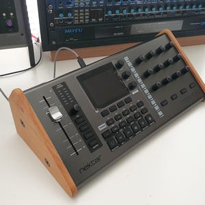 Puede incluir: Un controlador de teclado MIDI Nektar Impact LX25+ gris con un soporte de madera. El controlador tiene una pantalla grande, faders, perillas y botones. El teclado tiene 25 teclas.