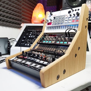 Puede incluir: Un soporte de madera con tres niveles para guardar sintetizadores de música electrónicos. El soporte está hecho de madera marrón claro y tiene un acabado de madera natural. Los sintetizadores son negros, blancos y grises. El soporte está diseñado para albergar un Korg Volca FM, un Korg Volca Keys y un Roland TB-03.