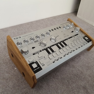 May include: A silver and black Roland TD-3 drum machine with wooden side panels. The machine has a variety of knobs and buttons, including a tempo knob, a track select knob, and a start/stop button. The machine also has a small keyboard with 16 keys.